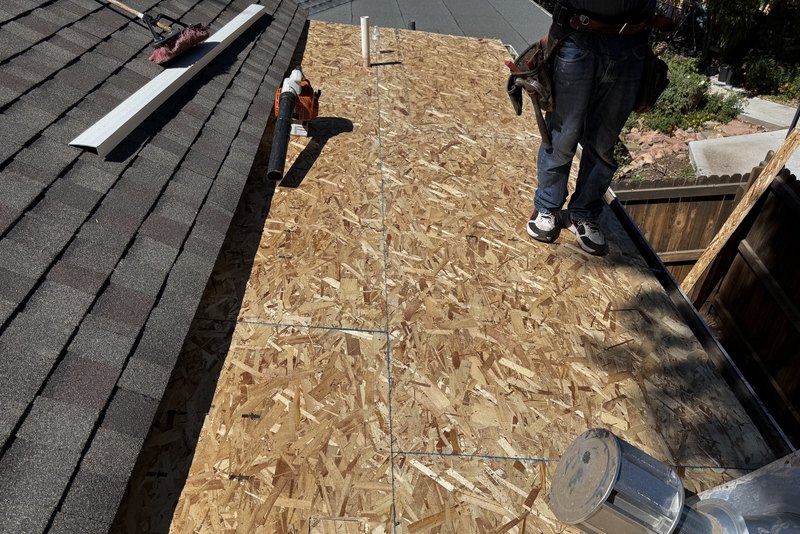 Exposed roof sheathing beside shingles and a worker’s legs