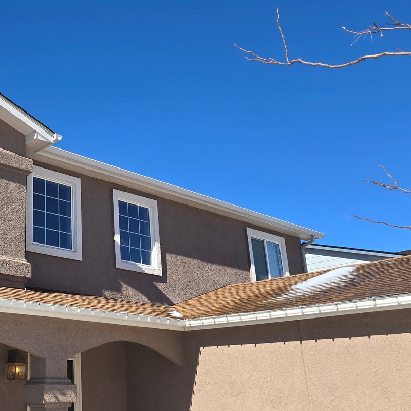 Snow on a lower shingle roof above white gutters on a two-story house
