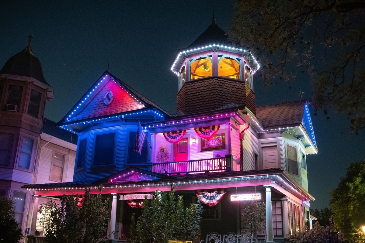 Victorian house at night with its roofline, turret, and porch outlined in blue, pink, and white lights