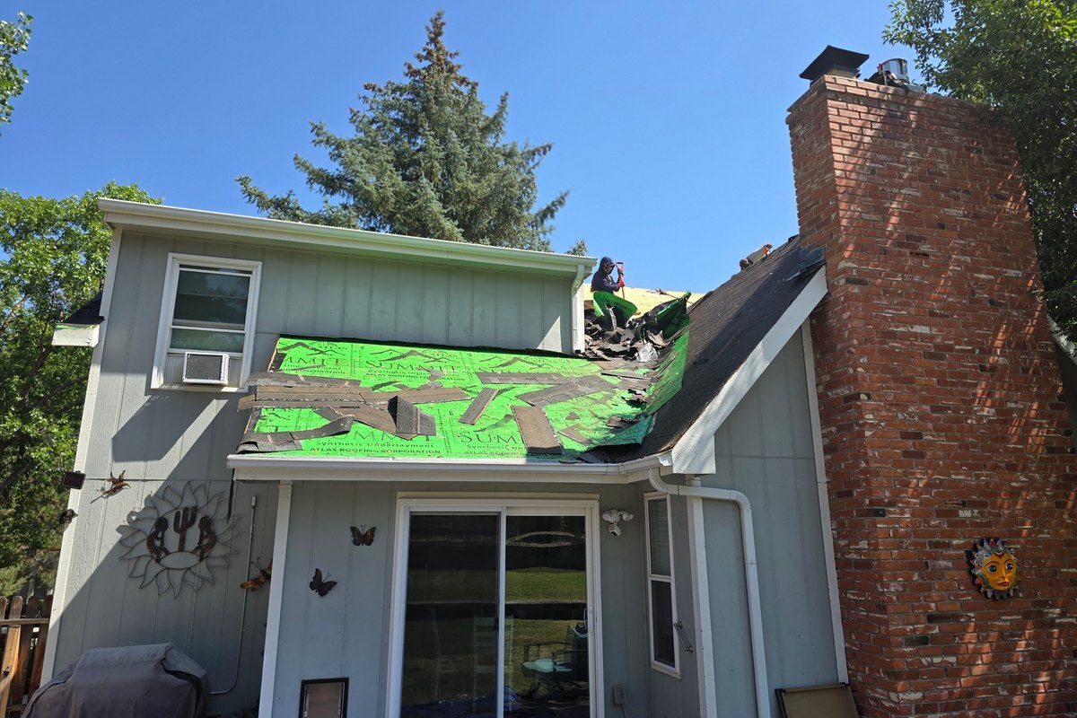 Partially stripped roof with green roofing underlayment beside a brick chimney