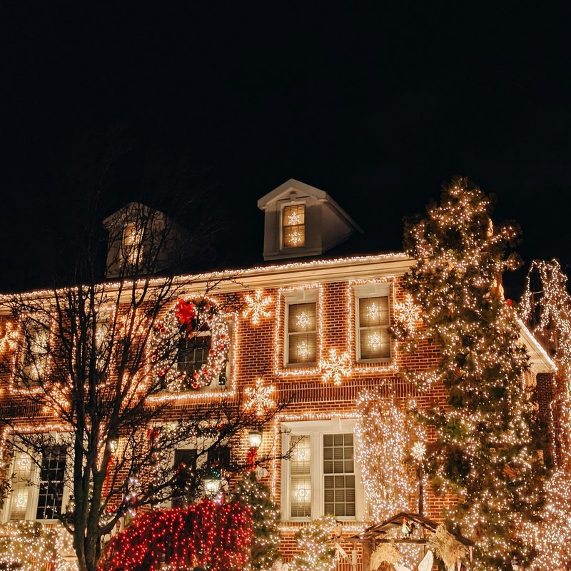 Two-story brick house at night with its roofline, windows, and front trees covered in warm white holiday lights